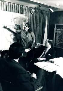 玛丽艾伦•韦尔奇(jack Welch)的照片,一个白色的女人,指着地图上看上去是一个社区。她与另外两个男人在一个房间里坐着看着她。