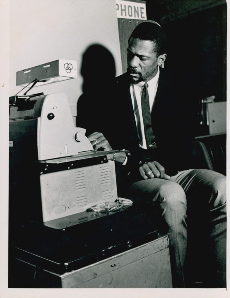 黑白图像的比尔-拉塞尔身穿一套黑色夹克和领带,坐在前面的一台机器。