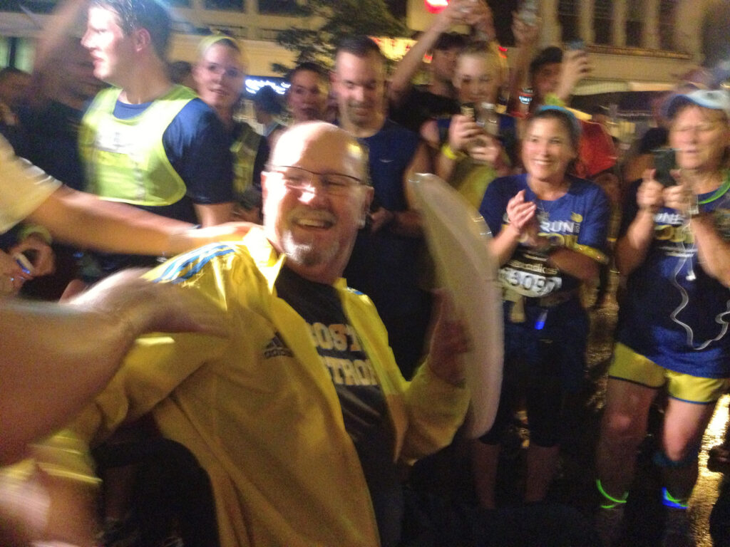 一个男人坐在轮椅上穿着黄色雨衣在波士顿强劲的衬衫是微笑和欢呼的跑步者穿一波士顿衬衫包围